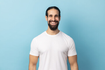 Portrait of bearded handsome man wearing white T-shirt standing looking at camera with satisfied face and smiling, expressing happiness. Indoor studio shot isolated on blue background.