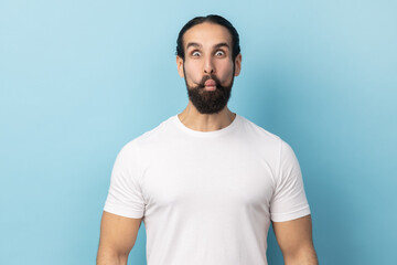 Portrait of childish man with beard wearing white T-shirt making fish face or sending kiss, having fun, kidding, pretending to be stupid. Indoor studio shot isolated on blue background.