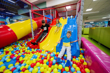 Happy kids playing at indoor play center playground. Children slides in colored slide into balls in...