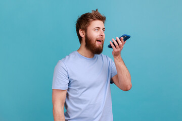 Portrait of positive optimistic bearded man recording voice message or talking to online assistant at his smartphone holding phone near mouth. Indoor studio shot isolated on blue background.