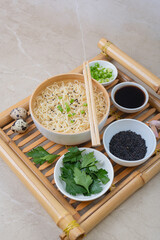 White bamboo plate (bowl) with egg noodles and plates with ingredients for it on a bamboo serving tray against a light background.