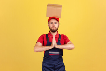 Relaxed courier man standing with cardboard box on his head, keeping hands in pray gesture, trying to calm down and resting during working day. Indoor studio shot isolated on yellow background.