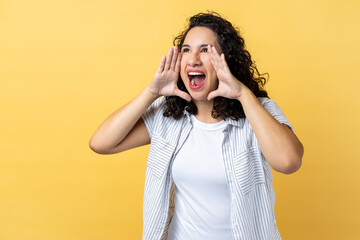 Portrait of attractive adorable woman with dark wavy hair opening mouth holding hands on face, screaming announcing her opinion. Indoor studio shot isolated on yellow background.