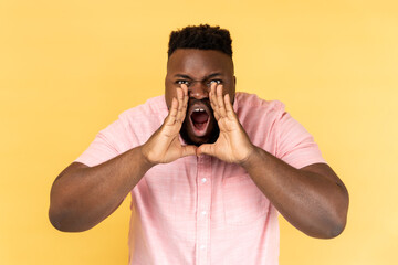 Portrait of angry aggressive man wearing pink shirt standing, holding arms near wide open mouth and screaming, trying to get attention. Indoor studio shot isolated on yellow background.