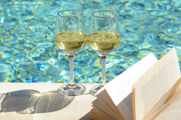 Glasses of tasty wine and open book on swimming pool edge