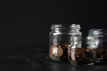 Glass jars with coins on black marble table, closeup. Space for text