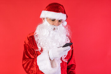 Man dressed as Santa Claus, consulting his mobile phone, on red background. Christmas, celebration, gifts, consumerism and happiness concept.
