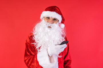 Man dressed as Santa Claus, consulting his mobile phone, on red background. Christmas, celebration, gifts, consumerism and happiness concept.