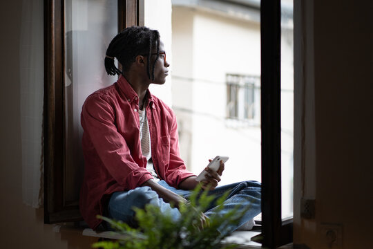 Pensive Dreamy Young African Man Sitting On Windowsill Holding Mobile Phone, Sad Black Student Guy With Smartphone In Hands Looking Through Out Open Window, Missing Friends While Studying Abroad