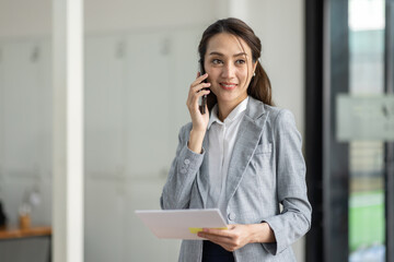 Young smiling Business asian woman talking on mobile phone, standing in workplace office with document in her hand