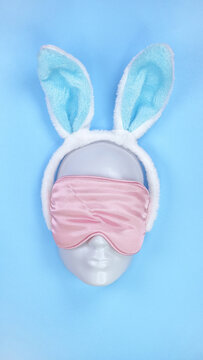Pink sleeping eye mask on mannequin face with blue fluffy Easter bunny ears on blue background, sleeping disorder. Holidays, Head accessory. Plastic face.