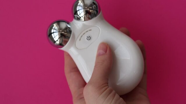 Microcurrent Facial Massager in woman's hands. Microcurrents to relax facial muscles from wrinkles on pink background.