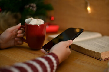 A female holding a cup of hot chocolate with marshmallows and her smartphone at her desk.
