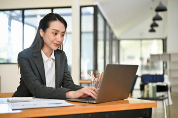 Professional Asian female accountant using laptop, typing on keyboard, working in her office.