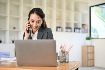 Fototapeta premium Asian female secretary talking on the phone with her business client while looking at laptop screen.