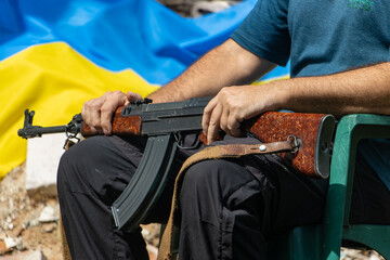 A man with an assault rifle sits in front of ruins with a Ukrainian flag