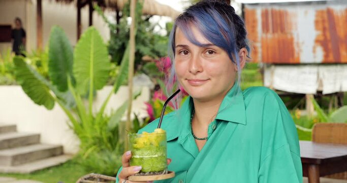 Young woman with dyed hair drinks cold green drink through straw, look straight to the camera. Tourist enjoying fresh cool beverage, sitting in a Balinese cafe. Then she smile and we see dental braces