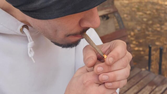 A handsome young mixed race guy with a beard and mustache lights a homemade cigarette using a lighter and exhaling thick tobacco smoke on a bench while sitting outdoors. Unhealthy Lifestyle