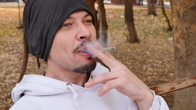 an adult young attractive guy smokes a cigarette with marijuana on a bench outdoors and enjoys exhaling the smoke. Mixed race man with dreadlocks holding a cigarette inhales tobacco smoke and enjoys