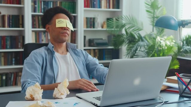 Young lazy African American man with funny paper stickers on eyes leans back in working chair trying to hide fatigue or drowsiness sits in office at table with laptop. Paperwork, freelancing