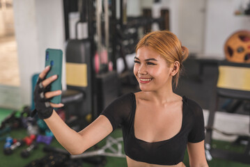 Fototapeta premium A happy asian woman poses while taking a selfie of herself during a training at the gym. Posting a workout photo to her social media.