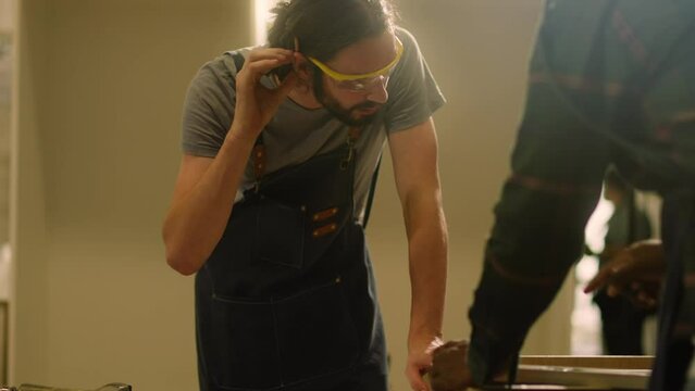 young hipster carpenter man tying his hair because his hair is blinding his eyes and making woodworks furniture in wood workshop.Carpenter SME owner at work measuring wooden by Cartridges Meters.