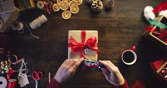 Hands, phone and box by worker wrapping christmas present in retail store at a table from above, night and coffee. Hands, employee and person taking picture on smartphone of festive gifts at a desk