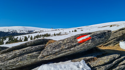 Directional path mark with Austrian flag painted on rock near Ladinger Spitze, Saualpe, Carinthia, Austria, Europe. Hiking trail on alpine meadow in Austrian Alps. Hills covered in snow, early spring