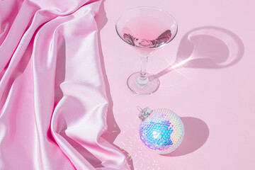 Creative layout with colorful Christmas baubles, cocktail glass and pink satin against a pink background. 80s or 90s retro fashion aesthetic concept. Minimal New Year or Christmas celebration idea.