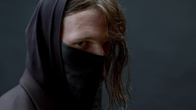 Horizontal close-up studio portrait of unrecognizable man with messy long hair wearing black outfit with mask on face standing in darkness
