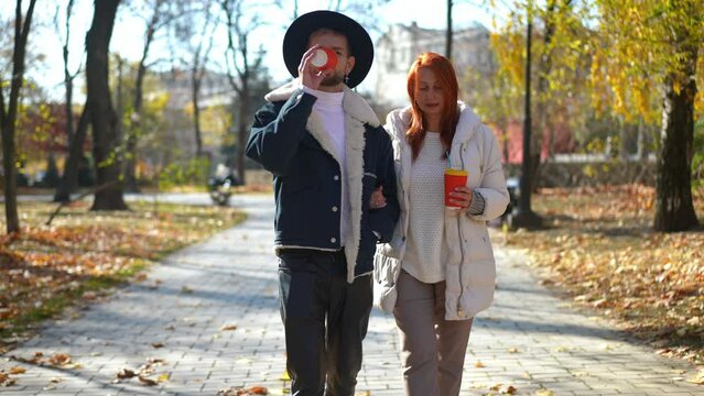 Front view confident Caucasian adult man and mature woman walking on park alley arm in arm talking. Dolly shot of LGBT son and redhead mother strolling chatting on autumn day outdoors