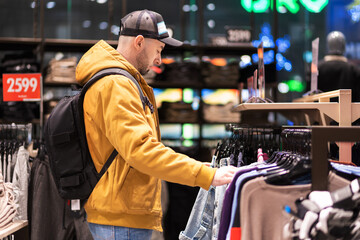Bearded Caucasian young man wearing backpack chooses clothes in store. Side view. Concept of...