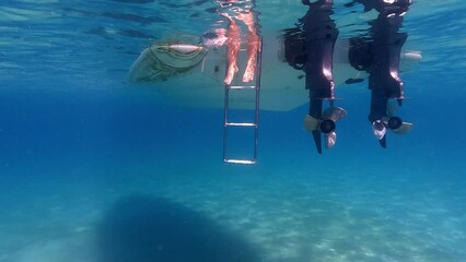 Slow motion under water tilt up shot of human legs and feet in seawater beneath surface protruding from double engine motorboat  ladder