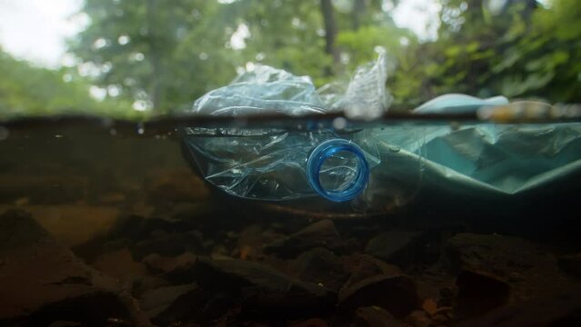 Used plastic bottle, medical mask and other rubbish lie on the surface of a forest stream in underwater view close up. Forest brook littered with various rubbish thrown out by irresponsible tourists
