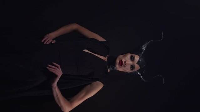 Vertical video An evil beautiful female witch-villain from a fairy tale with horns stands, playfully poses and seduces, looking into the camera in a dark black studio