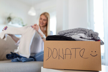Happy blond young woman sit on couch stuck clothes in donation box at home, caring biracial female volunteer put apparel in carton package, donate to needy people, reuse, recycle concept - Powered by Adobe