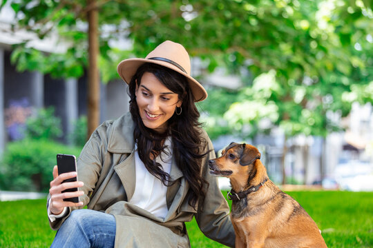 Portrait of pleased girl embracing funny dog and taking a selfie with her mobile phone. Smiling young woman in white shirt enjoying good day and posing with pet