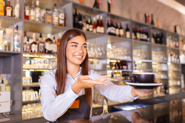 Beautiful female barista is holding a cup with hot coffee, looking at camera and smiling while standing near the bar counter in cafe