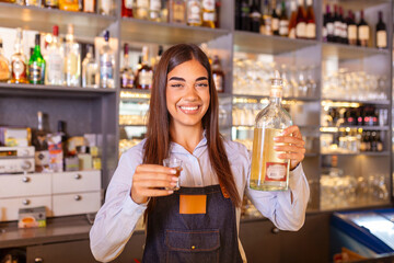 Beautiful female bartender is holding a shot glass with alcohol drink and a bottle in other hand, looking at camera and smiling while standing near the bar counter in cafe