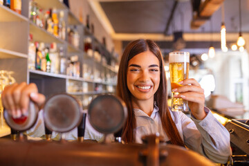 Beautiful smiling female Bartender serving a draft beer at the bar counter , shelves full of bottles with alcohol on the background