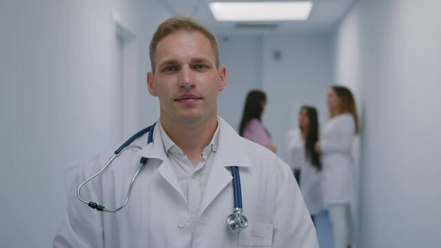 A male doctor in a white coat with a stethoscope looks into the camera against the background of colleagues in the clinic
