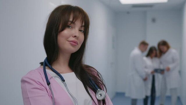The nurse looks into the camera and smiles standing in the corridor of the hospital against the background of working colleagues