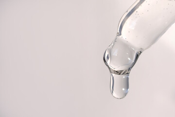 A drop of cosmetic facial gel or serum with air bubbles drips from a pipette on a gray background.