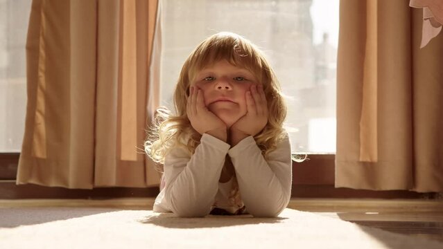 cute little blonde girl lie on carpet at home on floor and props her face with her hands, looks at camera with tired but funny look. Child toddler lies on floor on sunny day at home.