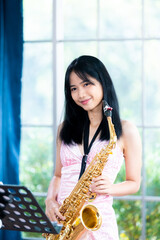 Young woman saxophonist with an instrument stands by the window. Cozy home interior. Concept of...