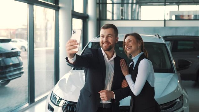 A Young Solid Couple Takes A Selfie On A Smartphone Against The Background Of A New Car In A Car Showroom