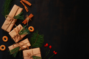 Christmas composition on a dark wooden background, top view. Several gift boxes wrapped in paper and tied with a rope, spruce branches, cinnamon, bagels and rose hips. Flat lay, copy space