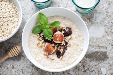 Oats porridge with fig fruits, dates, chopped nuts and honey in a white bowl, above view on a light-brown granite background