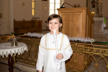 a handsome smiling boy in white clothes with a church candle stands in a Catholic church after...