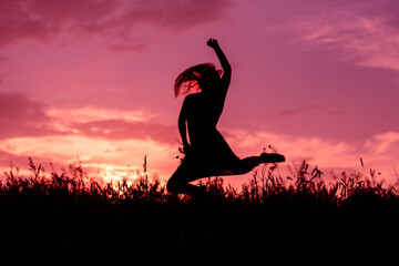 The silhouette of a jumping girl over the grass on the background of a pink sunset